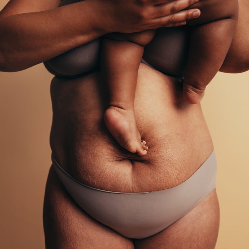 Midsection of mother carrying child while standing against brown background. Postpartum belly with stretch marks.